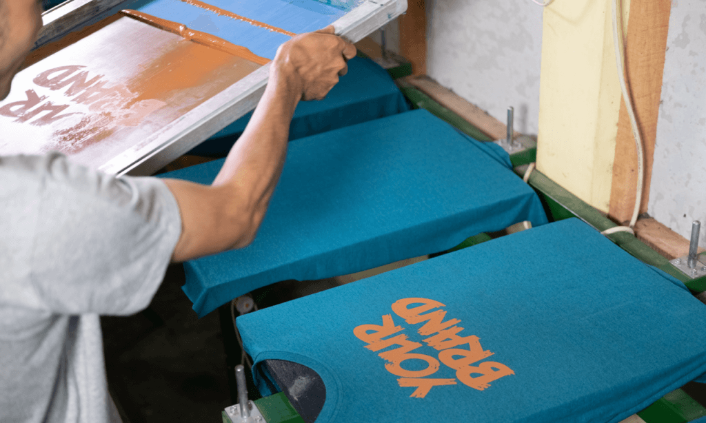 un uomo sta effettuato una stampa di magliette, applicando la scritta “your brand” in inchiostro arancione su una maglietta blu