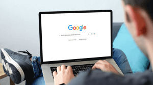 Uomo di spalle con un portatile sulla gambe. Sta navigando su Google, scrivendo nella barra di ricerca la query “dove personalizzare le magliette.”